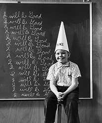 Boy wearing a dunce cap sitting in front of a blackboard